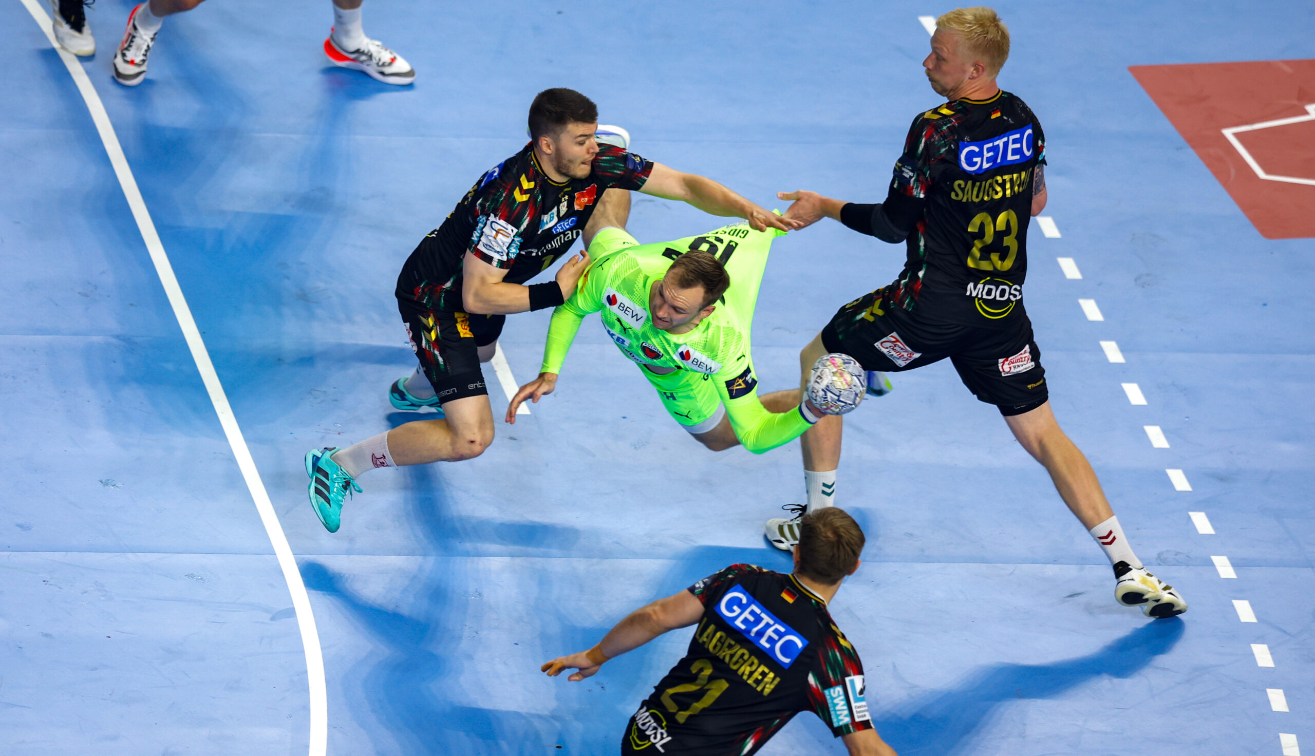 A player in a bright green jersey is being challenged by three opponents in black uniforms during an indoor handball game.