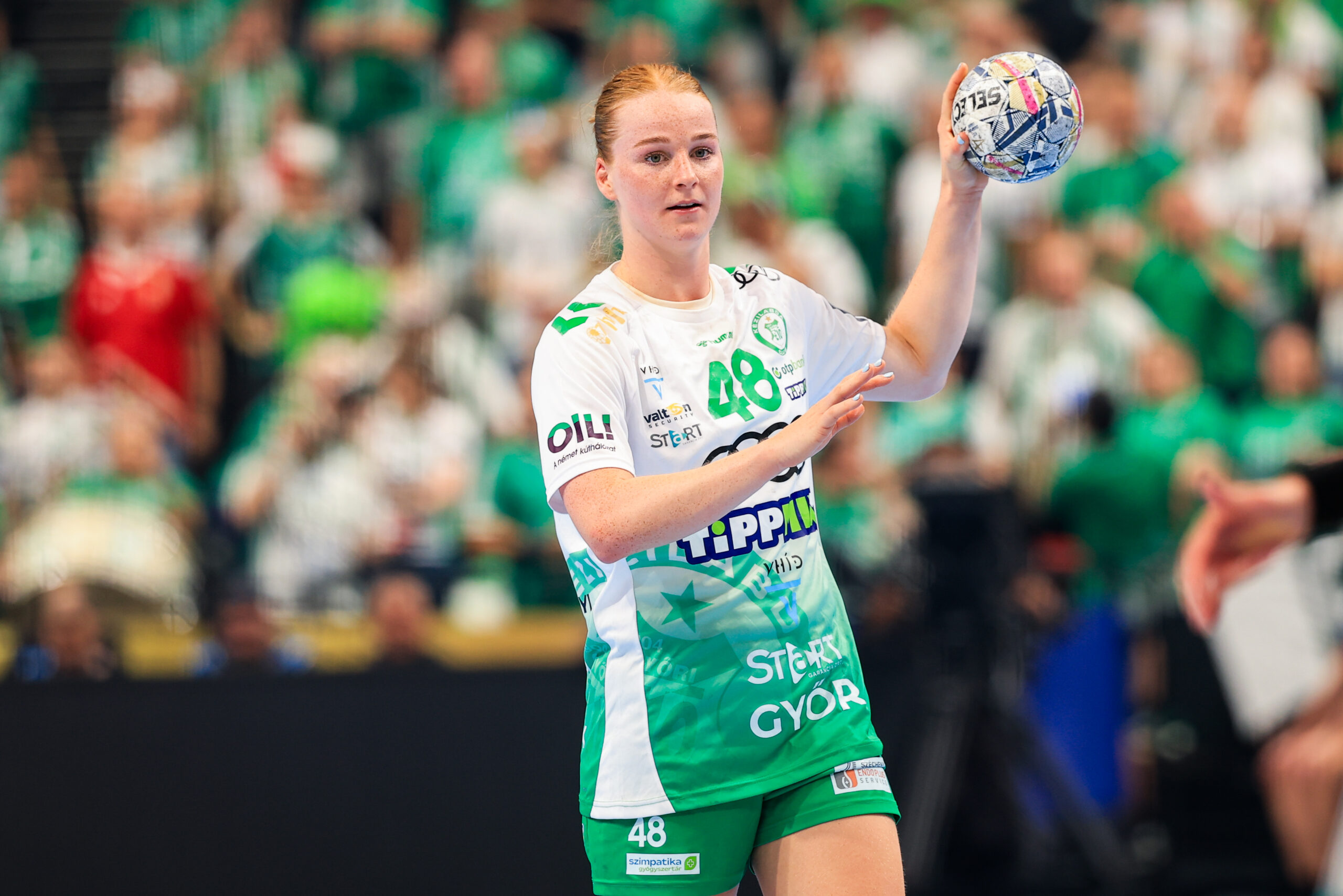 A handball player in a green and white uniform holds the ball above her head, preparing to throw, with spectators in the background.