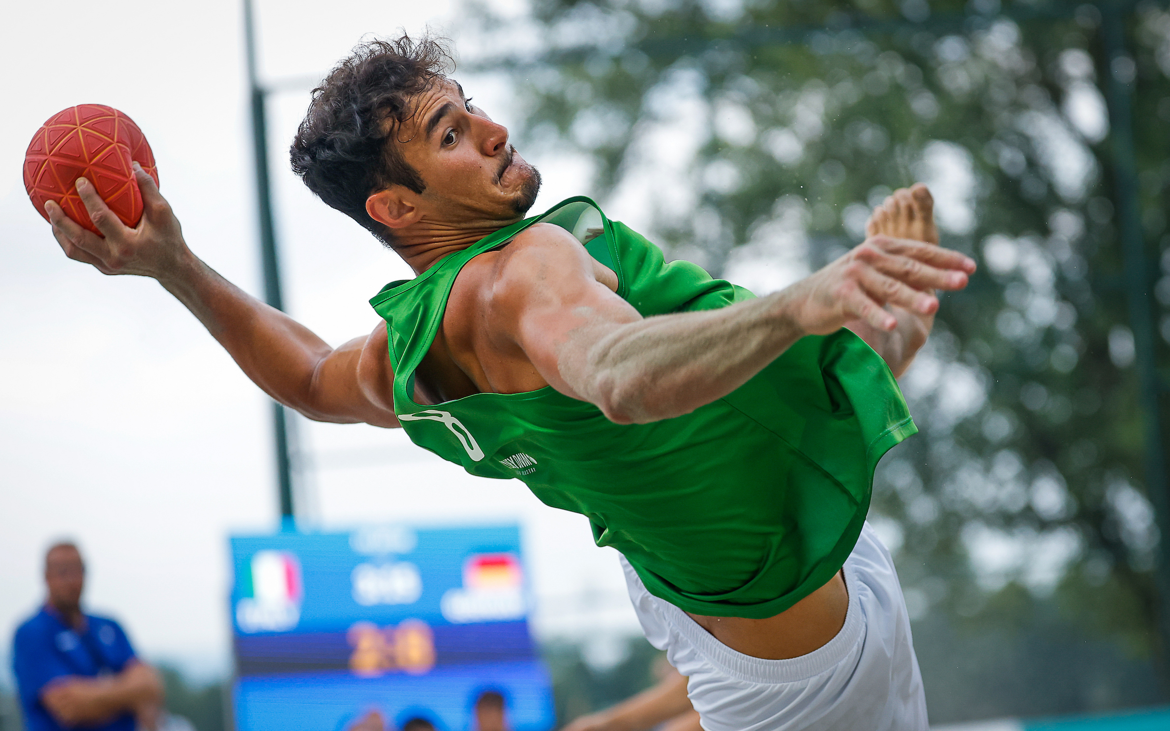 An athlete in a green jersey leaps to throw a red ball during an outdoor game.