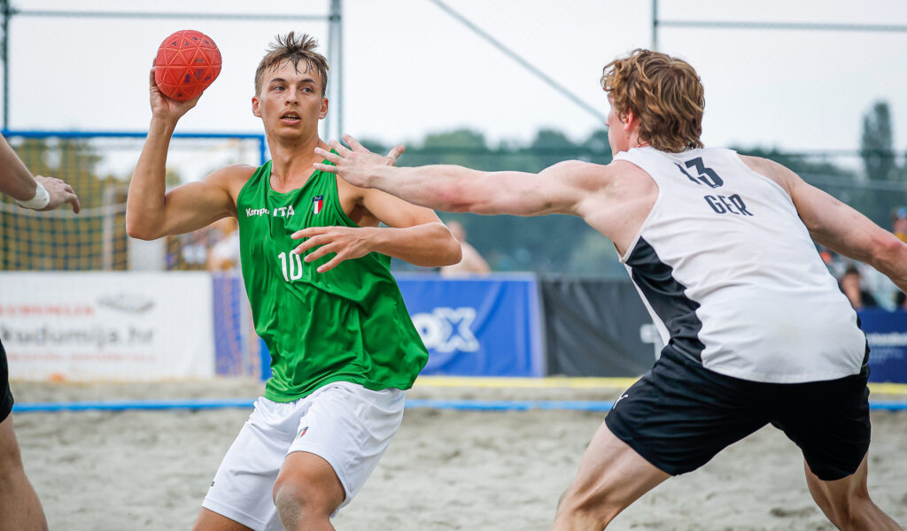 Two athletes compete on a sandy court, one in a green jersey preparing to throw a red ball while the other in a white jersey defends.