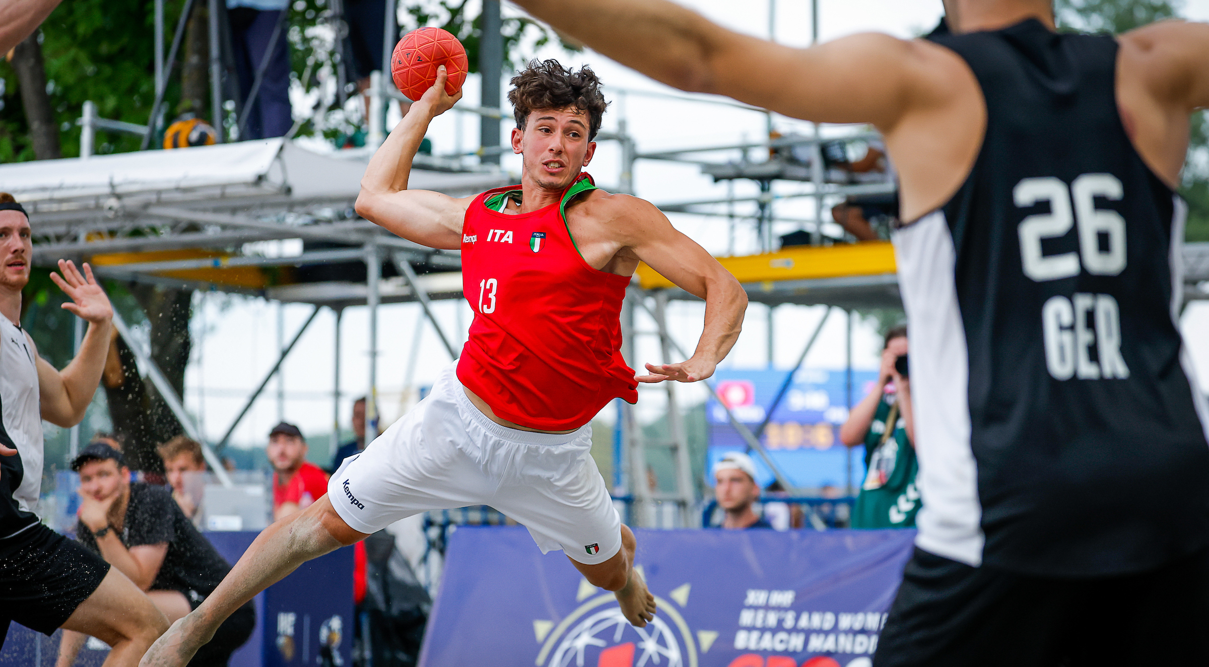 A player in a red jersey leaps to throw a beach handball while being defended by opponents on a sandy court.