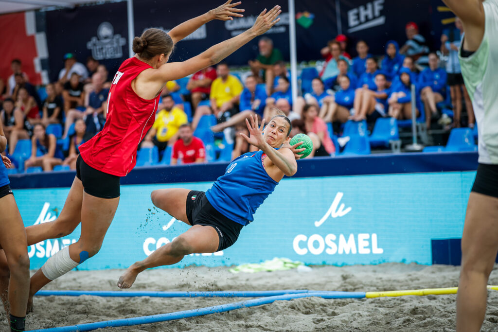 An athlete in a blue jersey dives to catch a ball while being defended by an opponent in a red jersey during a beach handball game.