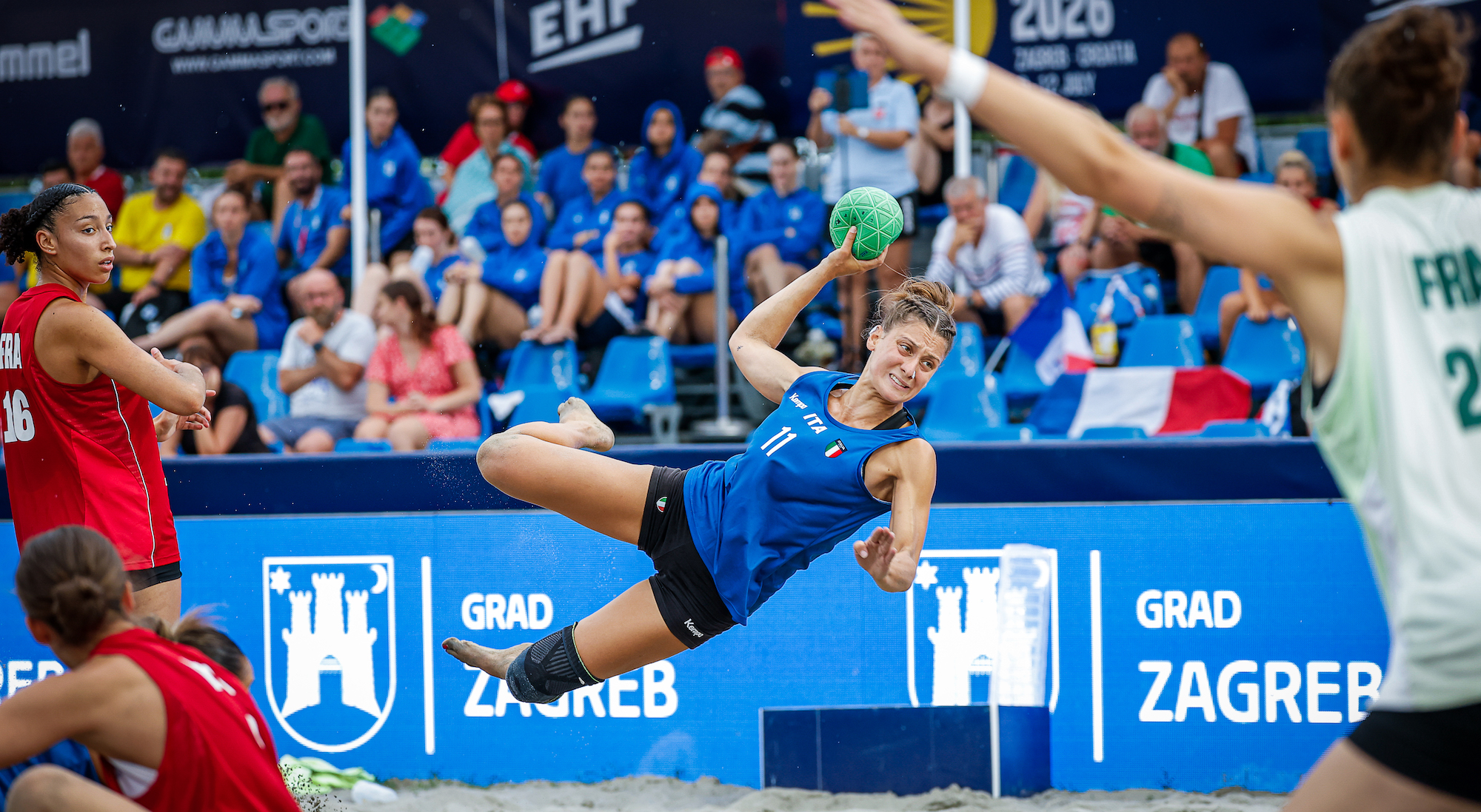 A player in a blue jersey leaps mid-air to catch a green ball during a beach handball match at the EHF Beach Handball Championship 2026 in Zagreb.