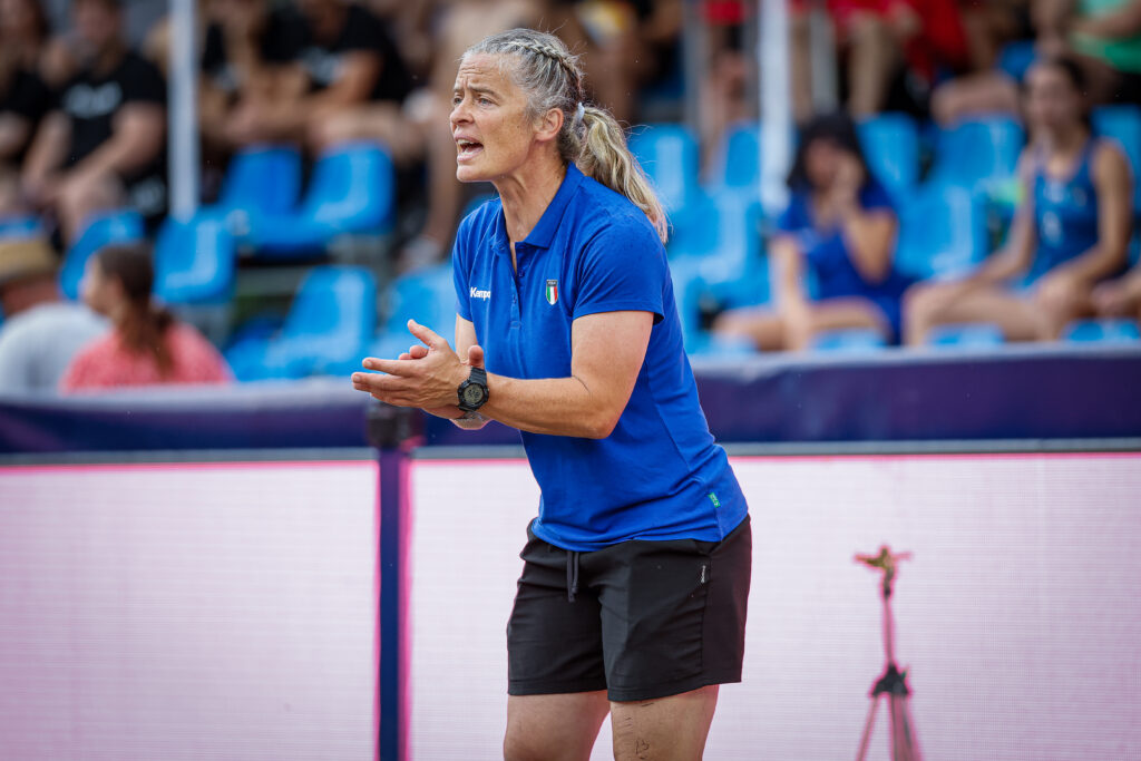 An athlete in a blue jersey and black shorts gestures animatedly while standing on a court, with spectators seated in the background.