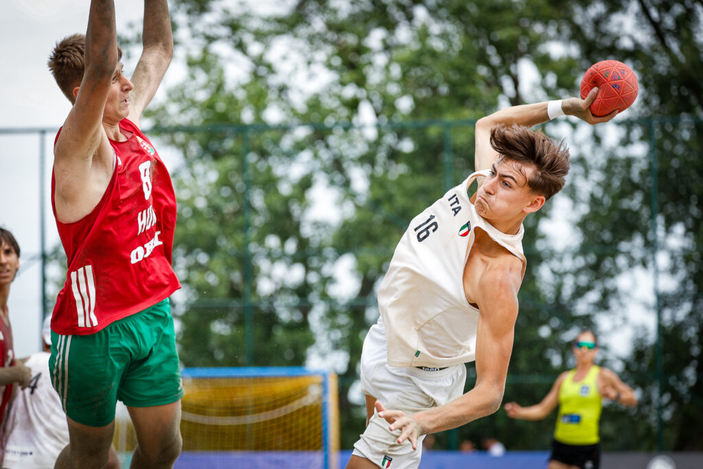 An athlete in a white jersey leaps to shoot a handball while being defended by an opponent in a red jersey.