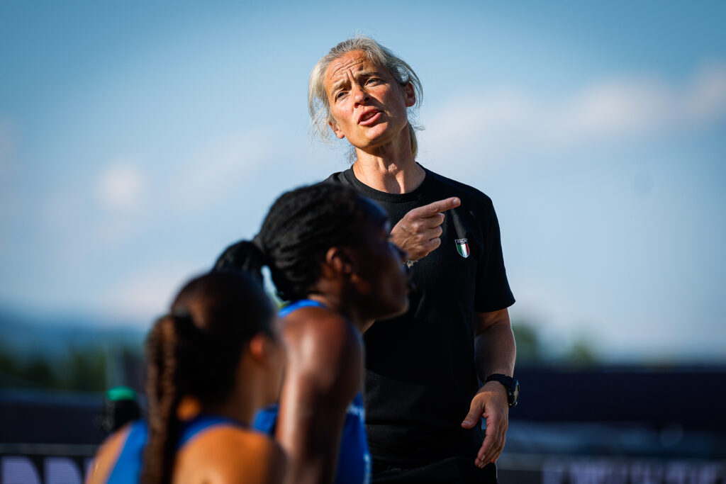 An athlete in a black shirt with a national emblem gestures while speaking to two other athletes during an outdoor training session.