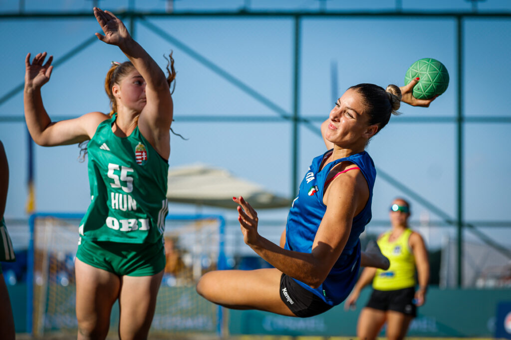 An athlete in a blue jersey leaps to throw a green ball while another player in a green jersey attempts to block.