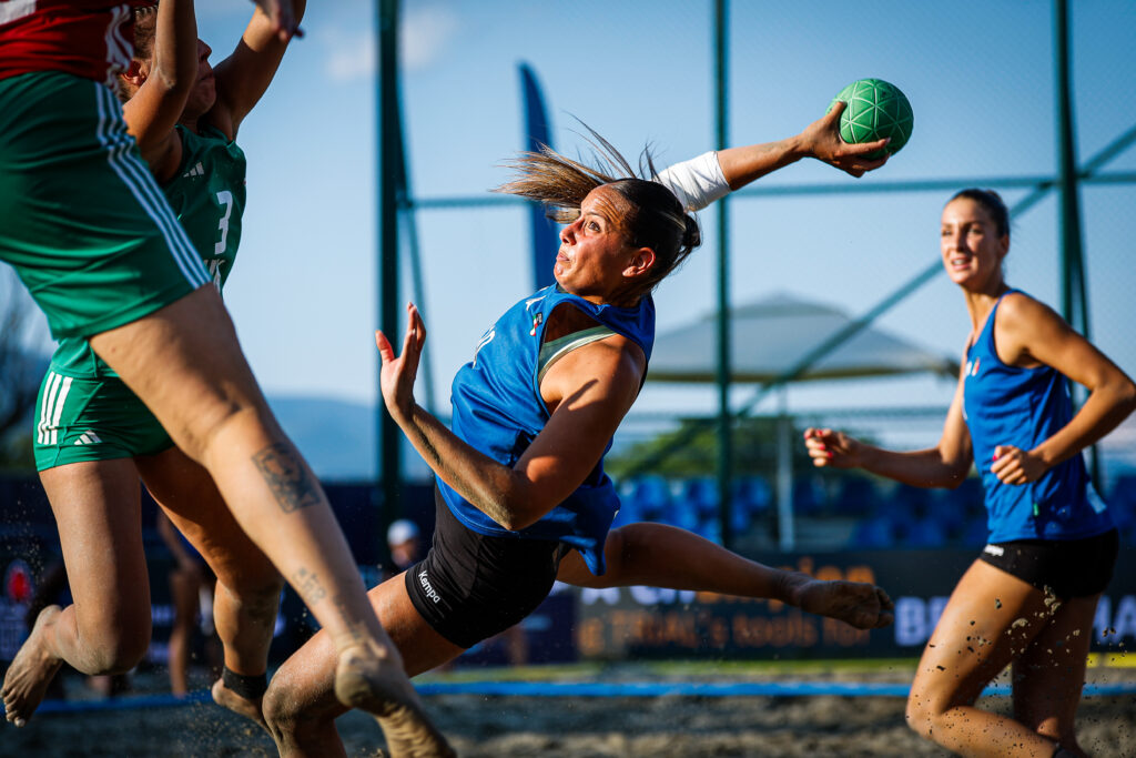 An athlete in a blue jersey leaps to throw a green ball during a dynamic beach handball match, with opponents and teammates in motion on the sandy field.