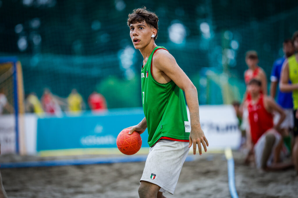 An athlete in a green jersey holds a red ball during a beach sport game on sandy terrain.