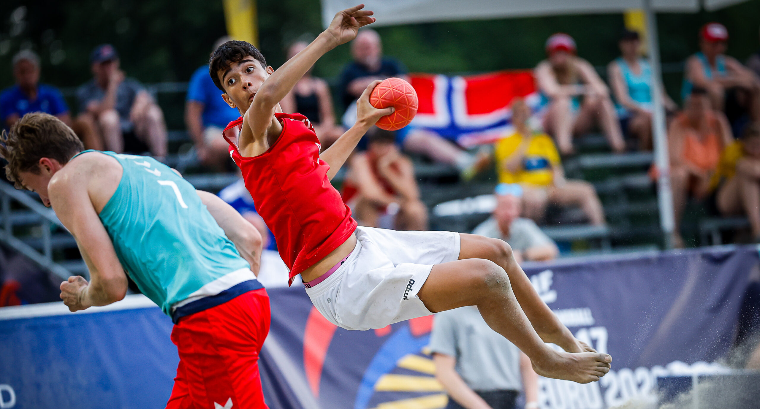 An athlete in a red jersey leaps through the air while holding a ball during a beach handball match, as another player in a teal jersey runs nearby.