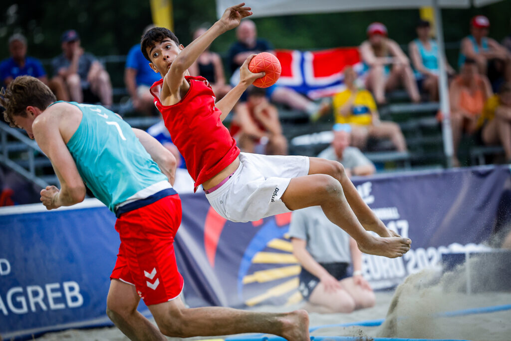 An athlete in a red jersey leaps through the air while holding a ball during a beach handball match, as another player in a teal jersey runs nearby.