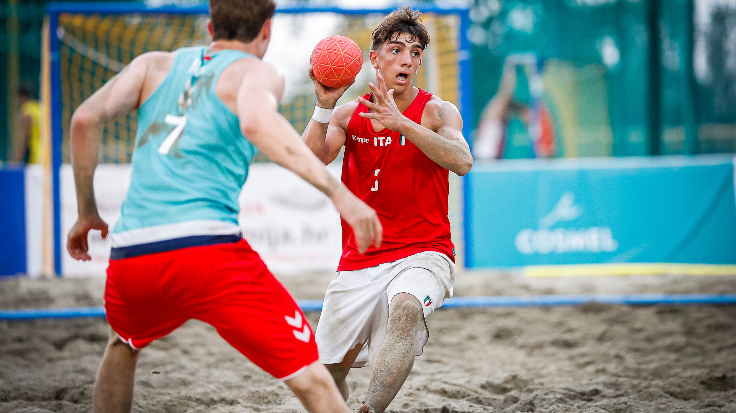 Two athletes compete in a beach handball match, with one in a red jersey holding the ball and another in a teal jersey defending.