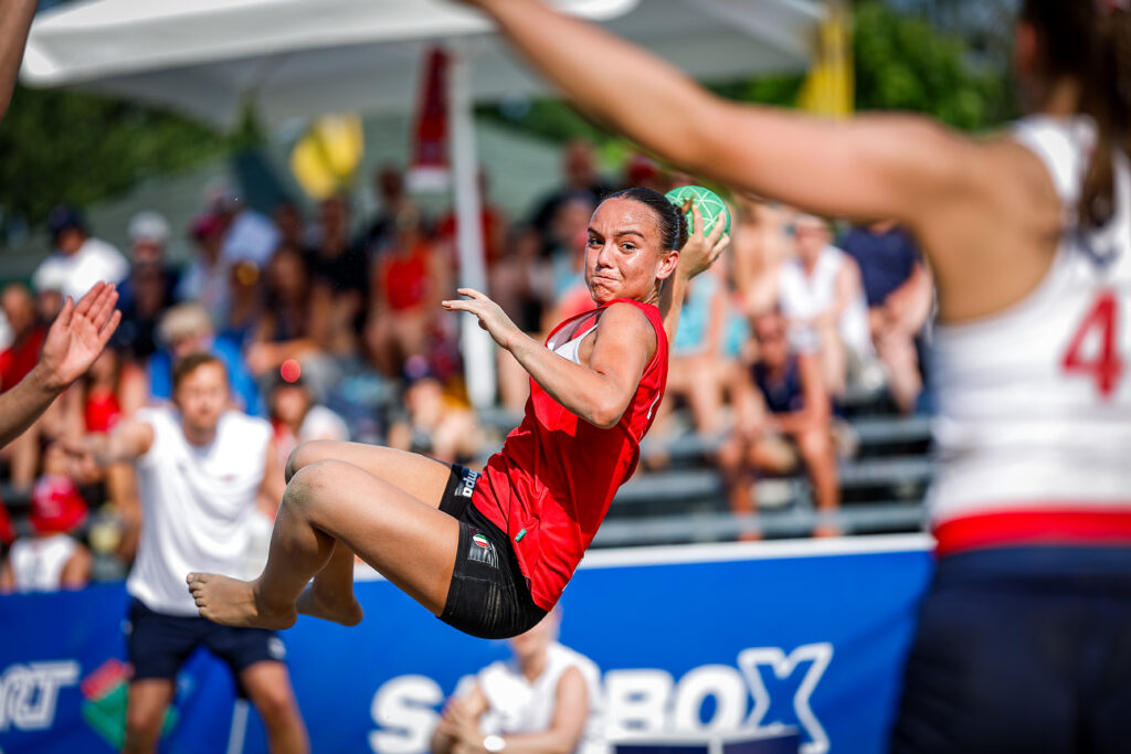 An athlete in a red jersey leaps mid-air to catch a green ball during a beach sport match, with spectators and teammates visible in the background.