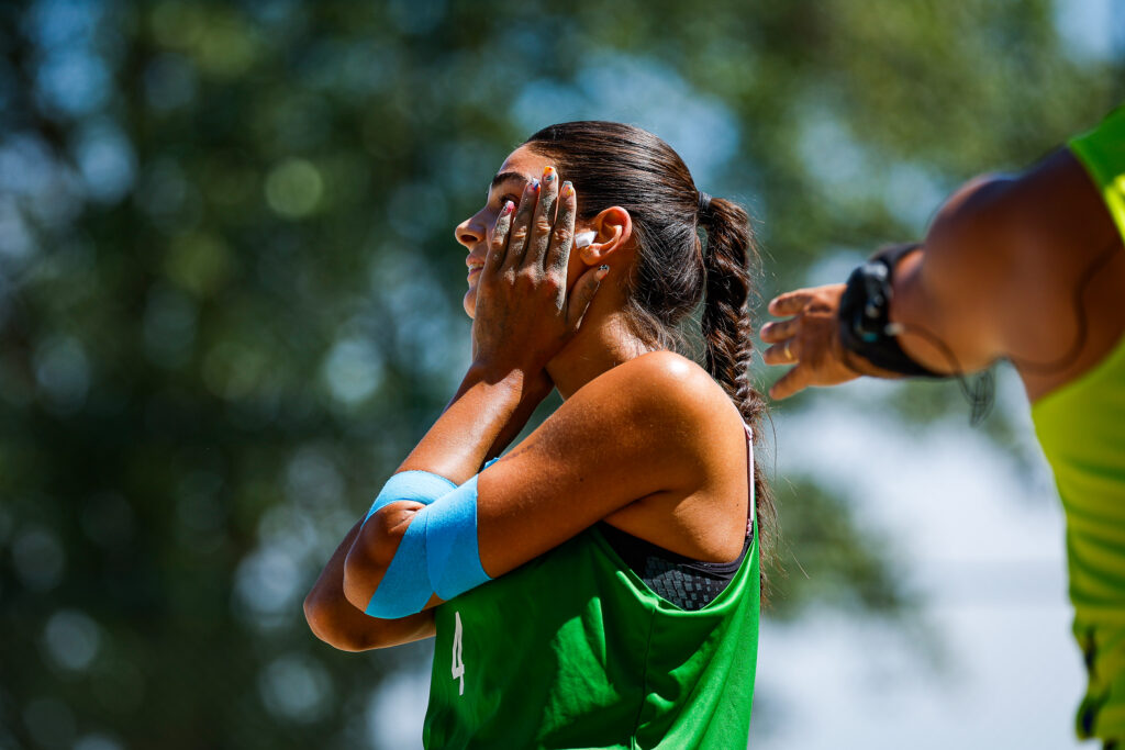An athlete in a green jersey rubs sunscreen onto her face while another person gestures nearby.